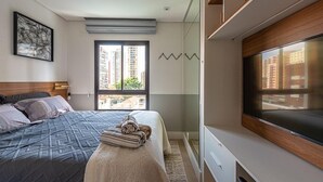 A modern bedroom with a large bed, a TV, and a view of tall buildings. - Condomínio Tag Decor (São Paulo)