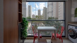 A balcony with a table and chairs, a washing machine, and a view of tall buildings. - Condomínio Tag Decor (São Paulo)