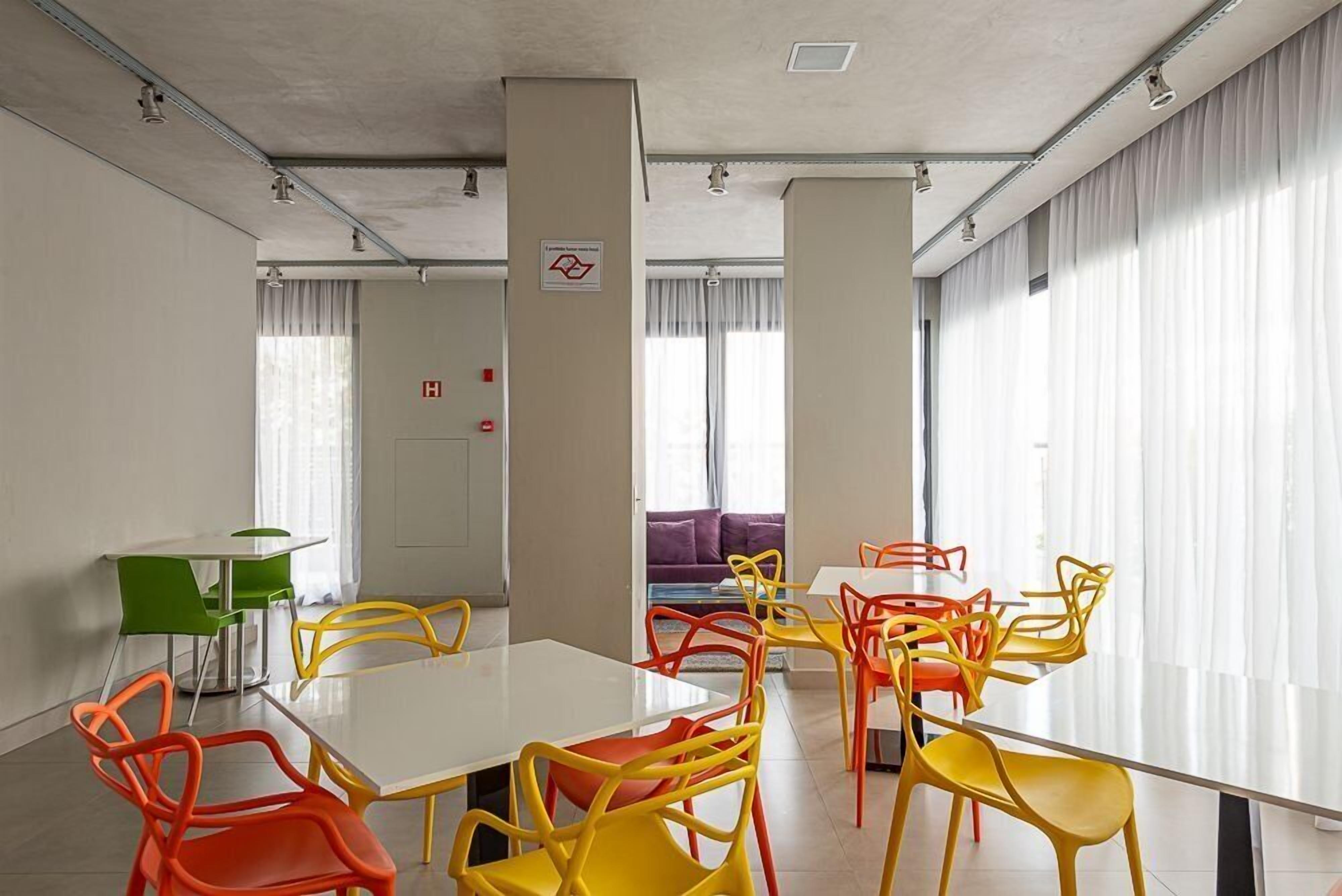 A conference room with a white table and colorful chairs, large windows with curtains, and a visible fire alarm.