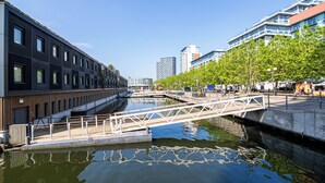 A modern urban canal with a pedestrian bridge, surrounded by contemporary buildings and trees. - 2-Bedroom Duplex Bliss steps from Excel London (London)