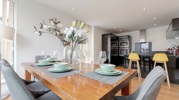 A modern dining area with a wooden table set for four, surrounded by grey upholstered chairs. - 2-Bedroom Duplex Bliss steps from Excel London (London)