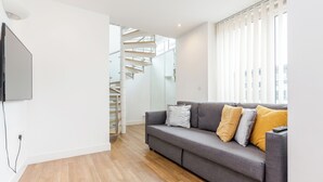 A modern living room with a grey sofa, wooden flooring, a flat-screen TV mounted on the wall, and a staircase leading to an upper level. - 2-Bedroom Duplex Bliss steps from Excel London (London)