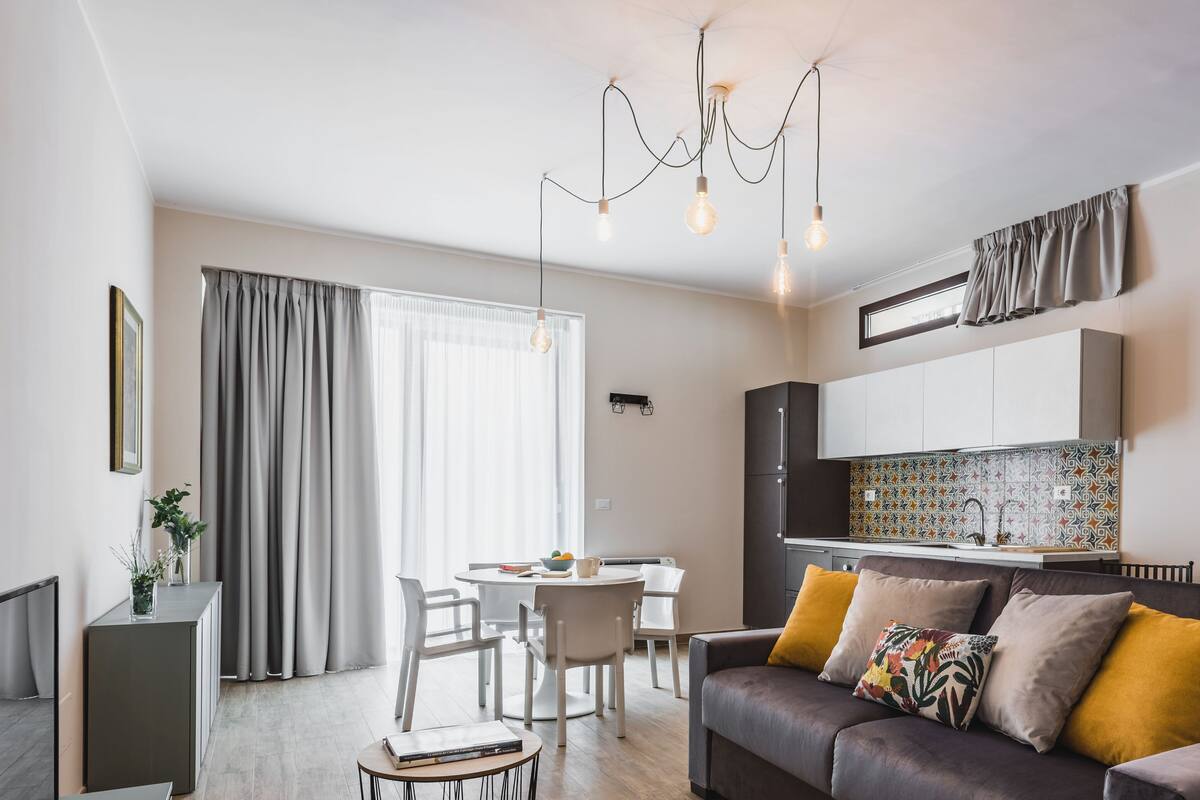 Een moderne woonkamer met een bank, eettafel en stoelen. Er is een keuken met kasten en een betegelde spatwand.