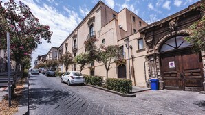 A cobblestone street lined with historic buildings, a white car parked on the side, and a blue trash bin near the entrance of a building with a large wooden door. - Venuto Homes by Wonderful Italy (Catania)