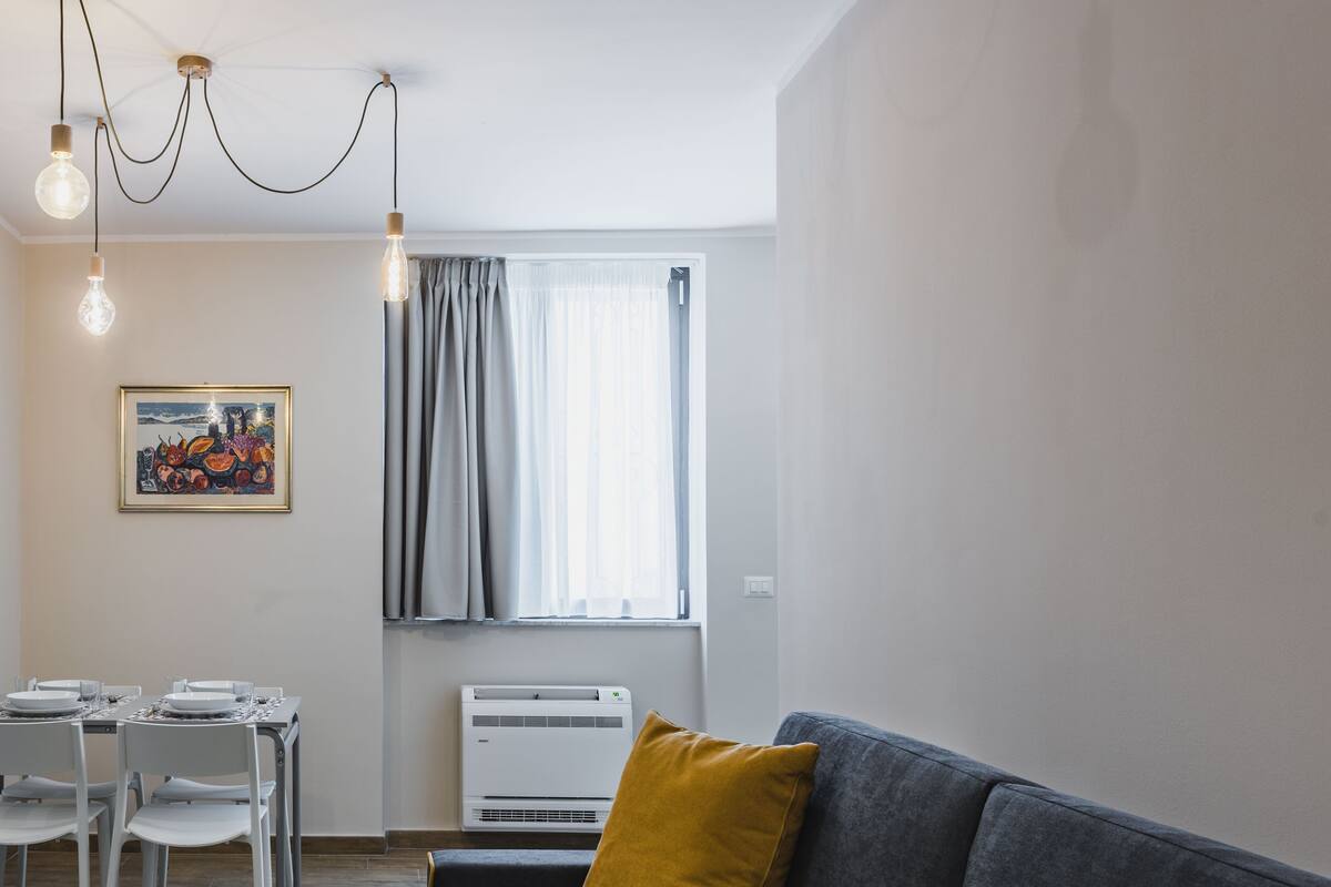 Een moderne woonkamer met een grijze bank, een eettafel met stoelen, een schilderij aan de muur en een raam met gordijnen.
