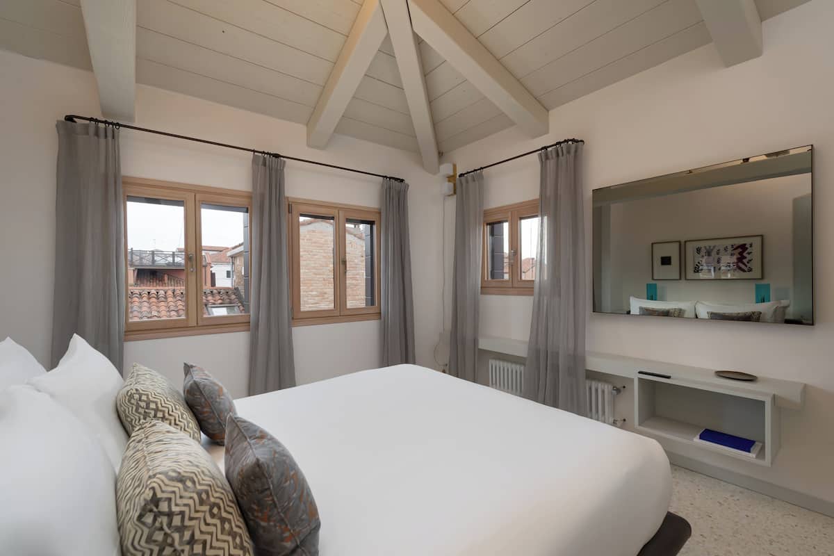 Ein Schlafzimmer mit einem großen Bett, grauen Vorhängen, einem weißen Nachttisch, einem Spiegel und Blick auf Gebäude.