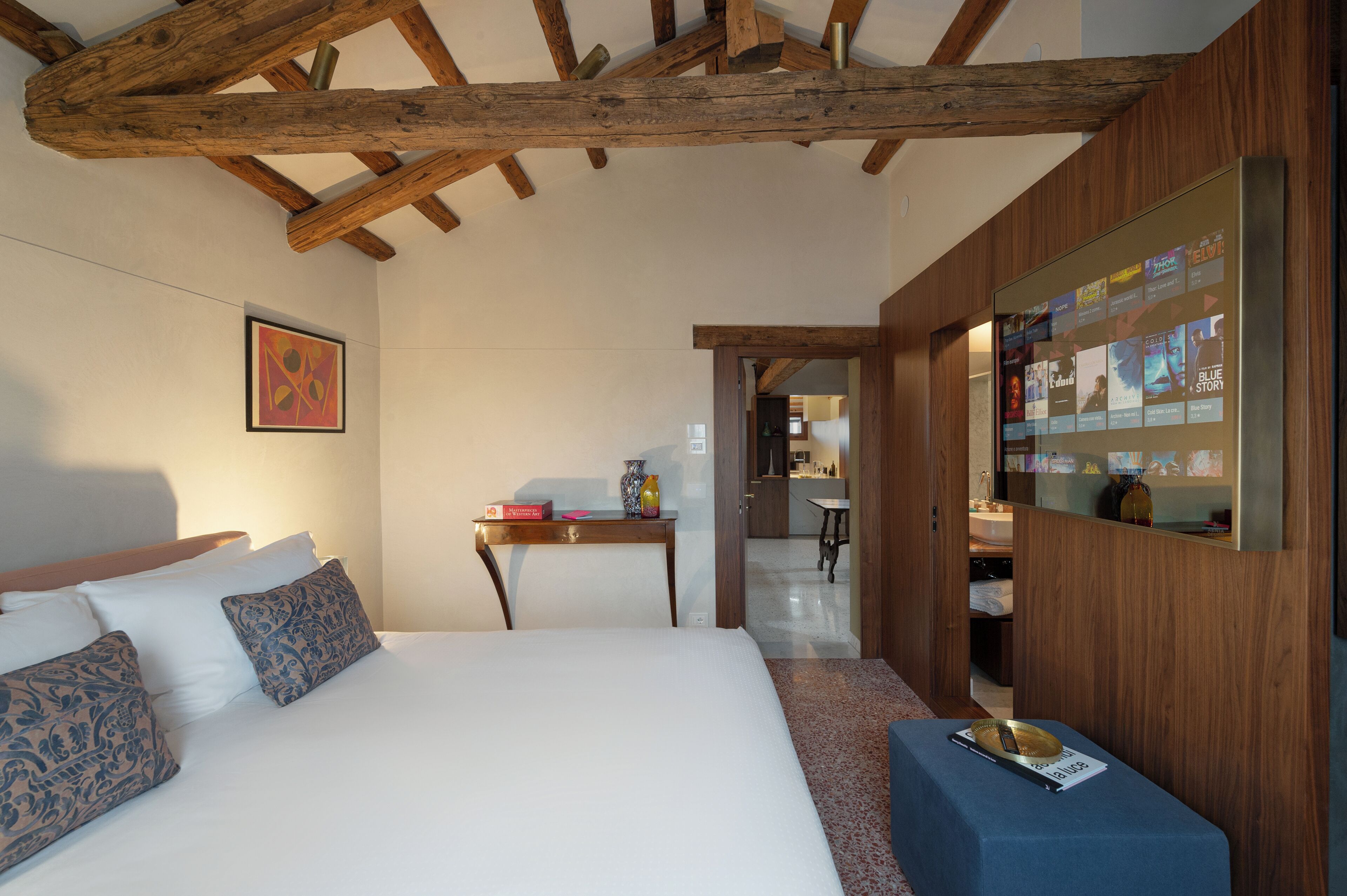 Un dormitorio con techo de madera, una cama con almohadas estampadas, una mesita de noche con un florero y una pantalla digital de pared.