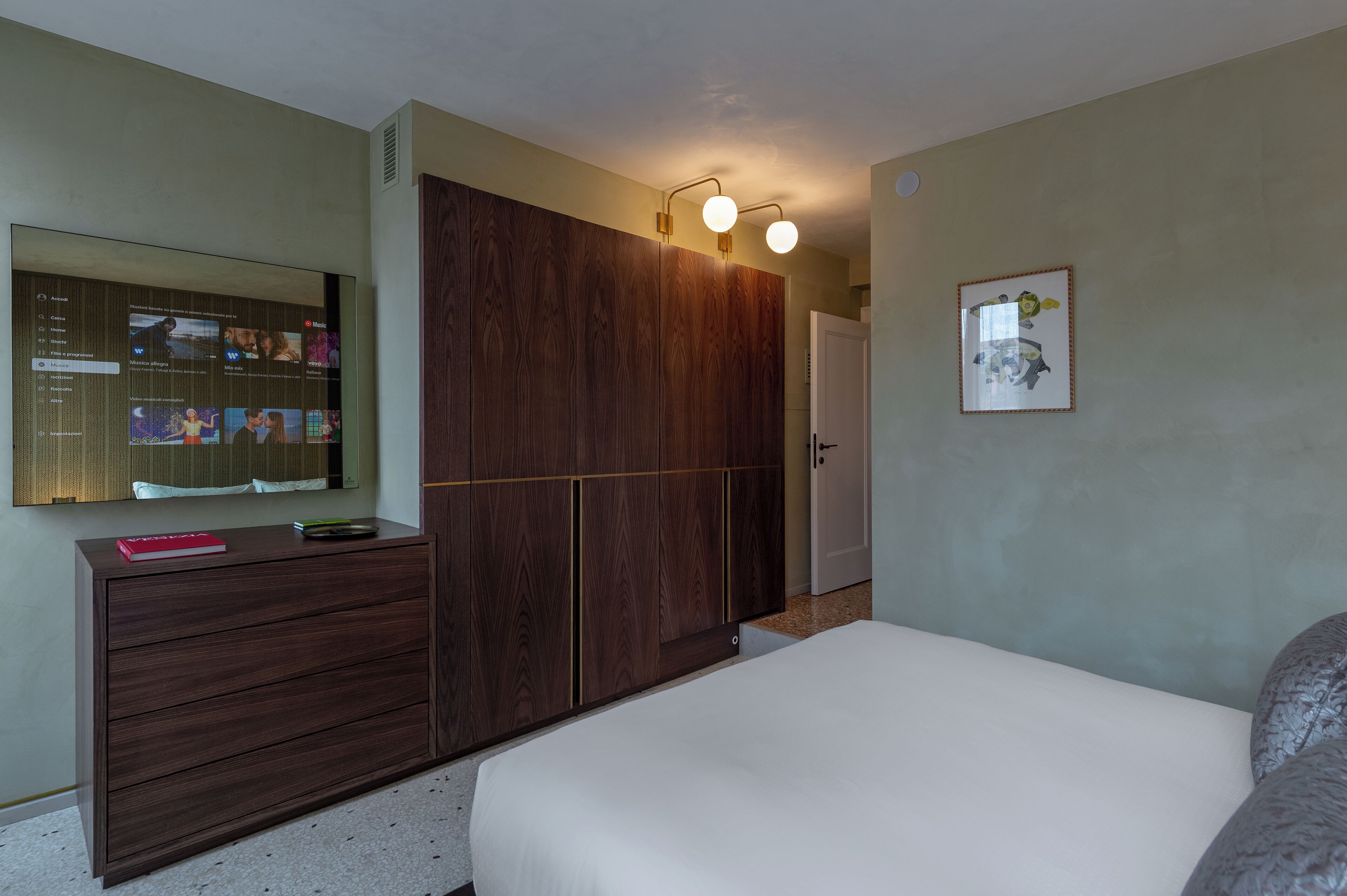 Ein Schlafzimmer mit einem großen Bett, einem Schminktisch, einem Kleiderschrank, einem Spiegel und einem an der Wand befestigten Fernseher, der verschiedene Sender zeigt.
