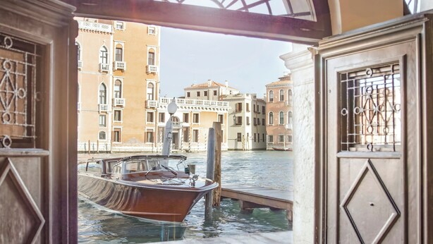 En trädörr som leder ut till en kanal med båtar, en utsikt över Venedigs byggnader och en dörrmatta med texten "Palazzo Garzoni Venezia".