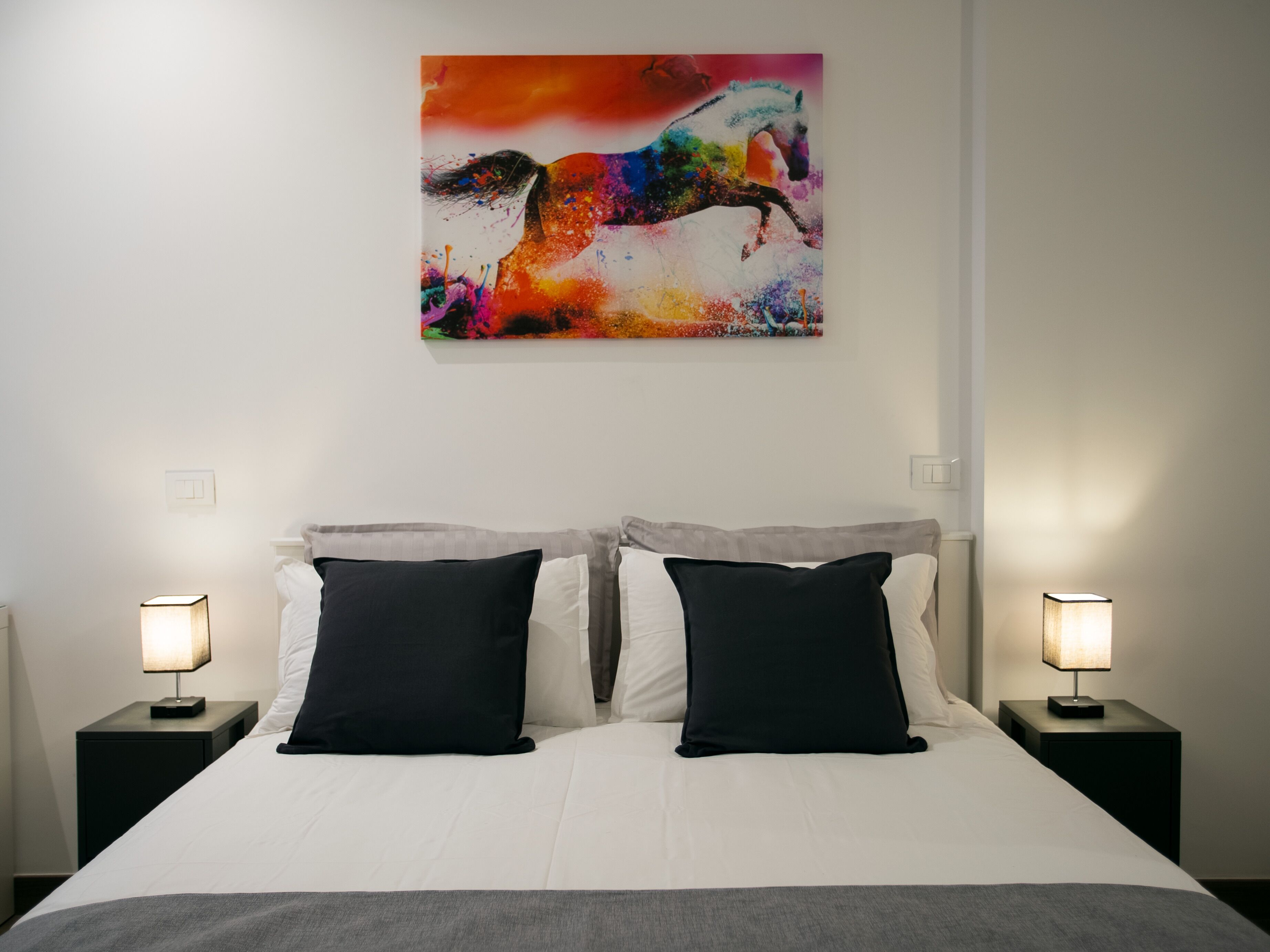 A hotel room with a bed, two black pillows, two bedside tables with lamps, and a colorful abstract painting on the wall.