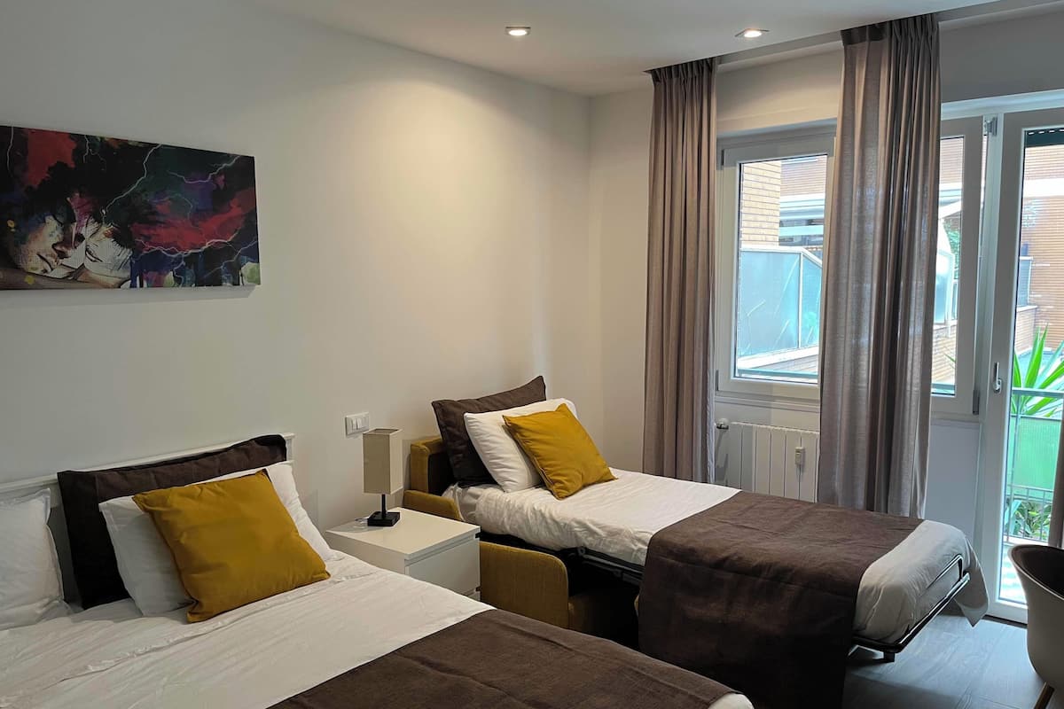 A hotel room with two beds, a nightstand, a lamp, and a large window with curtains.