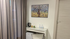 A compact room with a white desk, a small refrigerator, a chair, a painting of a tree, and a door. - Anastasio Vatican Rooms (Rome)