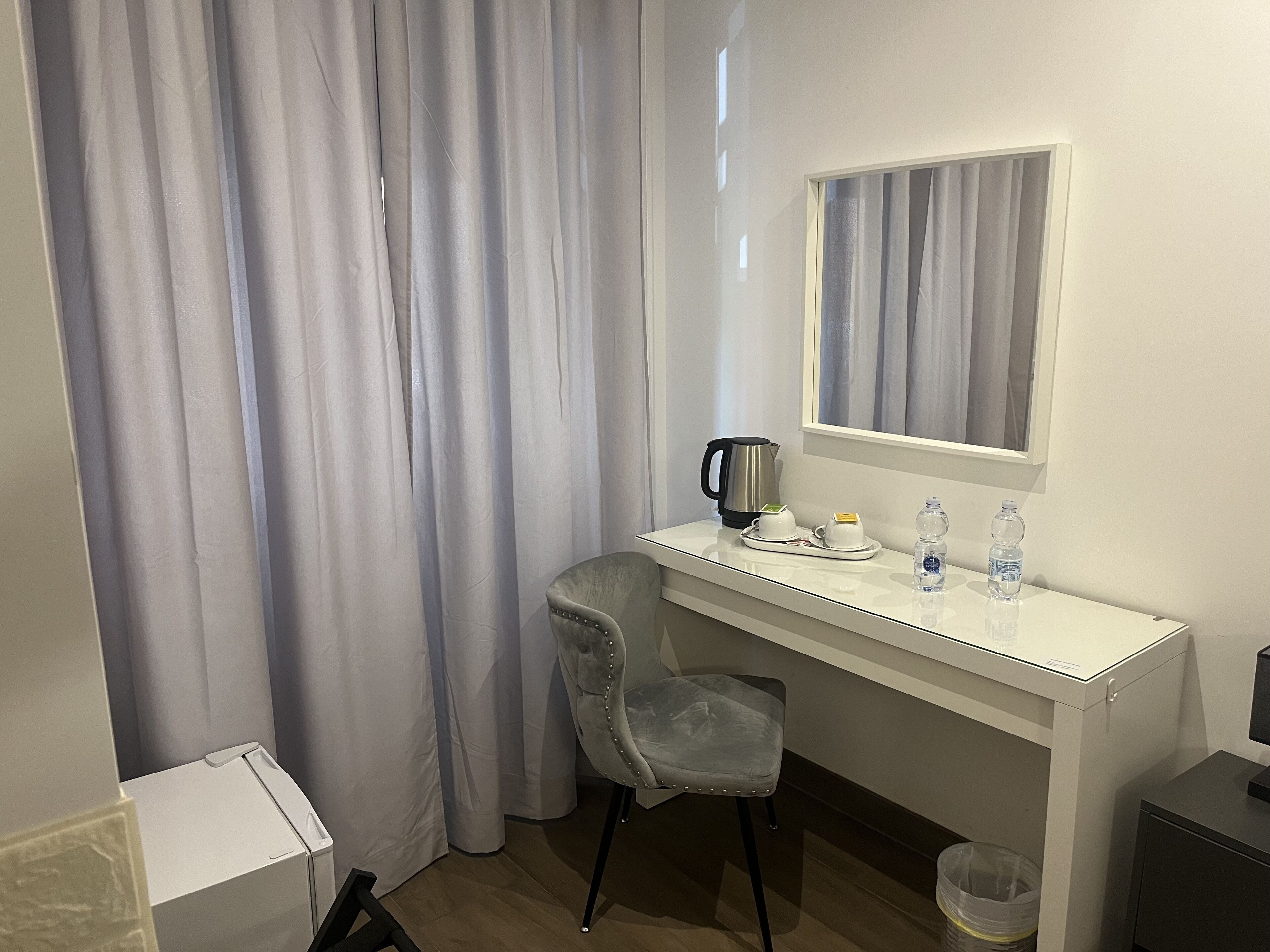 Habitación de hotel con un escritorio blanco, una silla gris, una tetera, dos tazas y dos botellas de agua.
