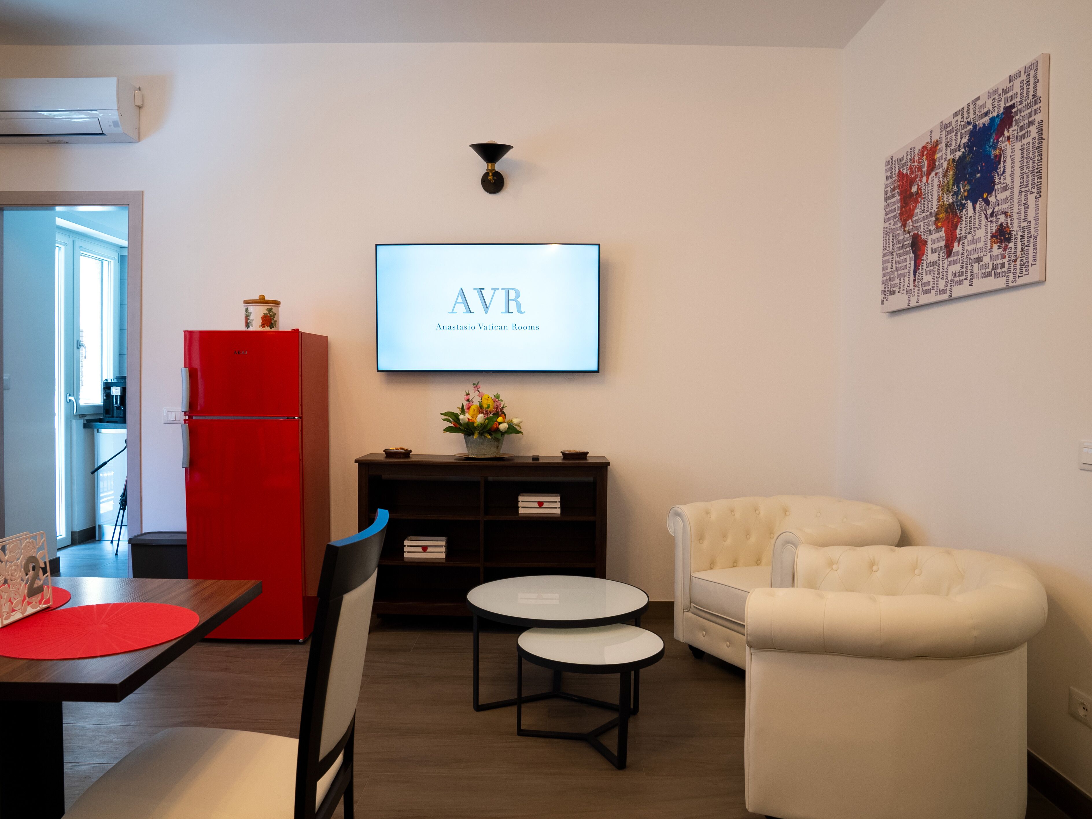 Een moderne woonkamer met een rode koelkast, een flatscreen televisie aan de muur, een kleine ronde tafel met twee stoelen en een witte fauteuil.