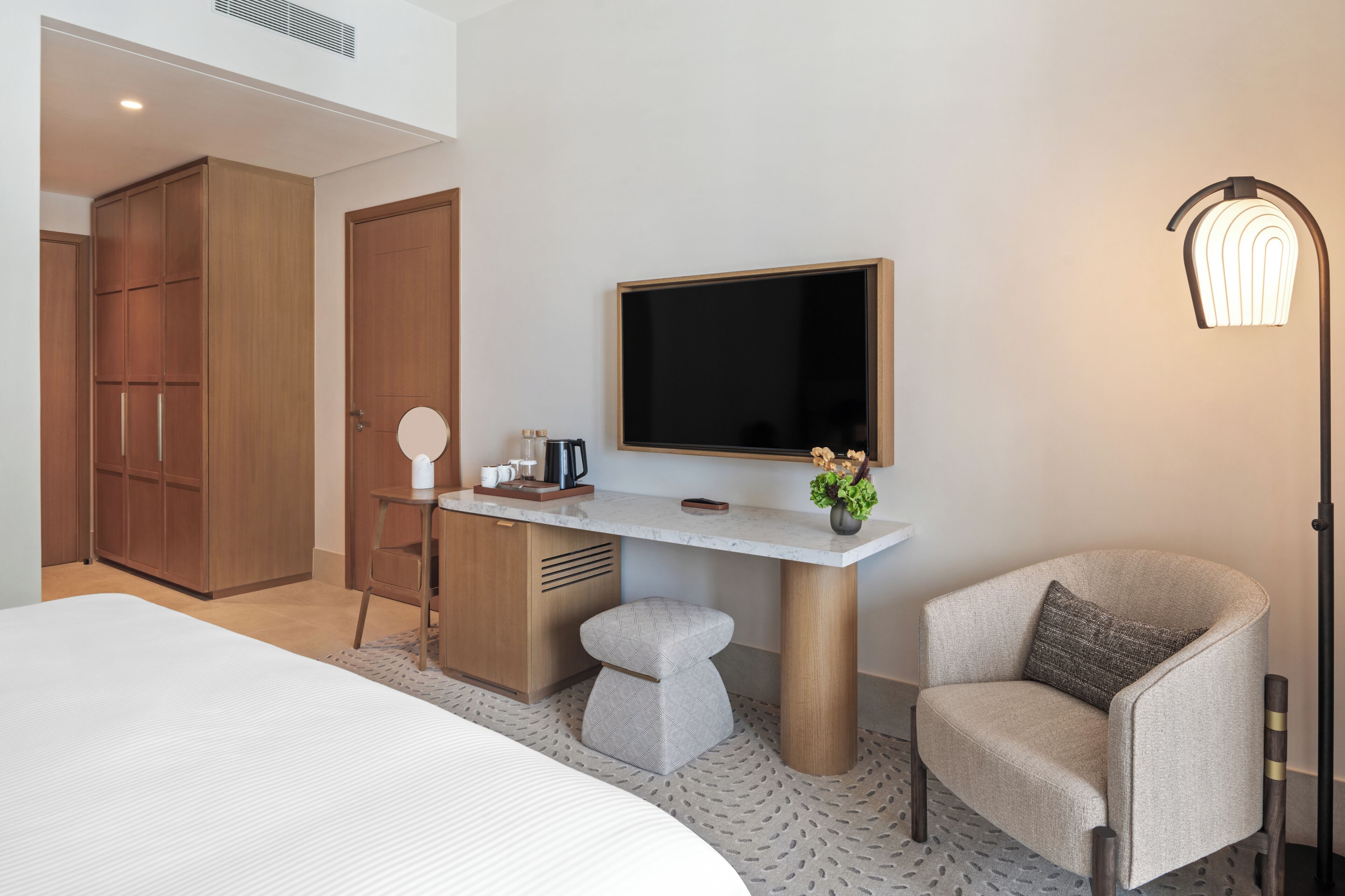A modern hotel room with a large bed, a desk with a flat-screen TV, a chair, and a wardrobe.