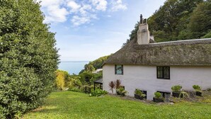 A white cottage with a thatched roof, surrounded by lush greenery and a view of the sea. - 5 Bed - Rose Cottage (Torquay)