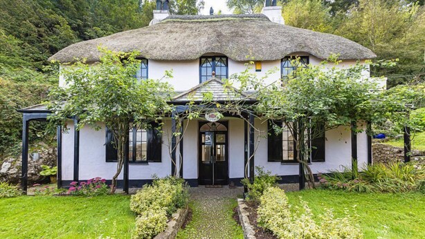 A white building with a thatched roof, surrounded by lush greenery and a cobblestone pathway leading to the entrance. - 5 Bed - Rose Cottage (Torquay)