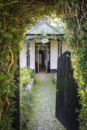 A pathway leading to a small, enclosed structure with a tiled roof, surrounded by lush greenery. - 5 Bed - Rose Cottage (Torquay)