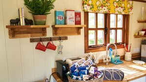 A kitchen with wooden shelves holding tea, coffee, and sugar boxes, a basket of snacks, and a window with a floral curtain. - Maple Shepherds Lodge (Dartmouth)