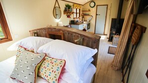 A cozy bedroom with a wooden headboard, a bed with white linens, and two decorative pillows. A small kitchenette is visible in the background. - Maple Shepherds Lodge (Dartmouth)