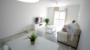 A modern living room with a white sofa, a dining table with chairs, a TV on a white cabinet, and a standing fan. - 135 Livingtarifa Luna (Tarifa)