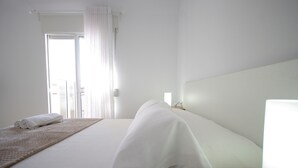 A hotel room with a bed, a bedside table, a nightstand, a window with curtains, and a view of the outside. - 135 Livingtarifa Luna (Tarifa)
