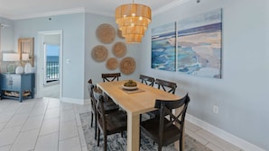 A dining area with a wooden table and chairs, a hanging light fixture, and a coastal landscape painting on the wall.