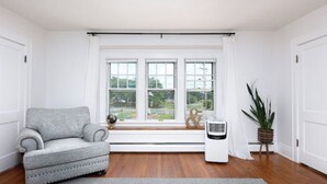 Ein Zimmer mit grauem Sessel, einem Standheizgerät, einem Blumentopf und einem Fenster mit Blick auf Grünflächen.