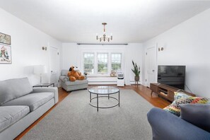 Ein geräumiges Wohnzimmer mit grauem Sofa, rundem Couchtisch, grauem Sessel, Fernseher und einem großen Fenster mit Tageslicht.