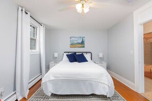 Ein Schlafzimmer mit einem Bett, einem Deckenventilator, einem Nachttisch, einem Bild an der Wand und einem Fenster mit Vorhängen.
