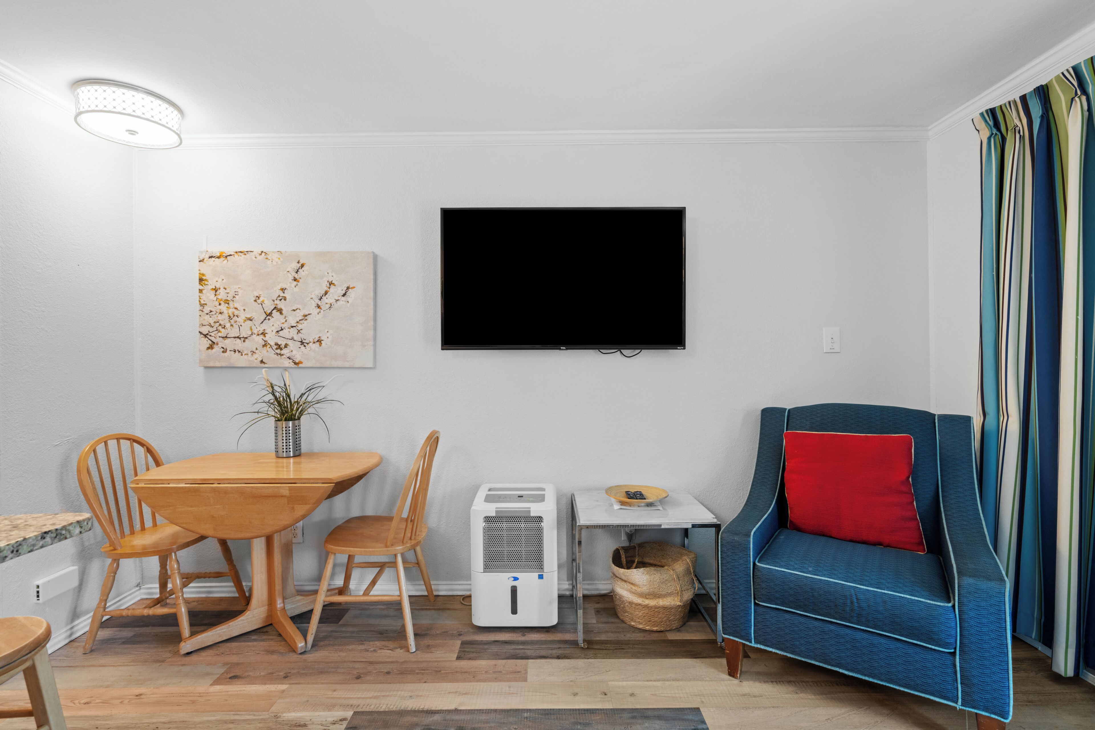 A modern living room with a flat-screen TV, wooden dining table, chairs, a blue sofa with a red pillow, and a small table with a plant.