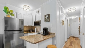 A kitchen with white cabinets, a stainless steel refrigerator, a granite countertop, wooden stools, and a ceiling light fixture. - Beach Front- Cozy for Family In Casa Del Mar (Galveston)
