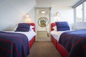 Une chambre avec deux lits simples, chacun doté d’un cadre rouge et d’une couette à carreaux bleus. On y trouve deux lampes de chevet, un miroir sur le mur et une fenêtre sur le côté droit.
