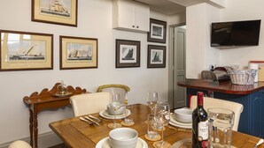 A dining area with a wooden table set for four, framed nautical paintings on the wall, a television mounted above the counter, and a bottle of wine on the table. - 3 Bed Cove Cottage (Torquay)