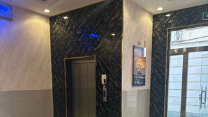 A modern elevator with a marble wall and a digital control panel, a glass door entrance, and a green artificial grass area visible through the door.