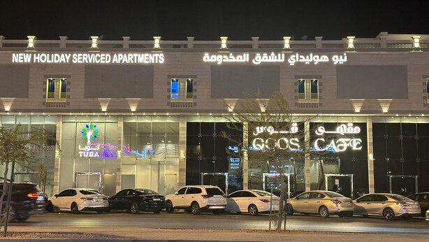 A hotel with a sign for "New Holiday Serviced Apartments" and Arabic text. There are several cars parked in front.