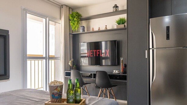 A modern hotel room with a bed, a desk with a TV displaying 'NETFLIX', a refrigerator, and a balcony with a view. - Estúdio Moderno com AC e Rooftop (São Paulo)