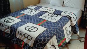 A bed with a Gucci-patterned bedspread and pillows, a tiled floor, and a plain wall. - Mary queen hotels and suites  (Ekpoma)