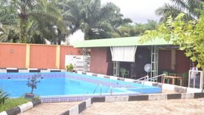 A swimming pool with a tiled border, a covered seating area, and a building with a green roof. - Mary queen hotels and suites  (Ekpoma)