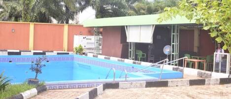 Una piscina con borde de azulejos, una zona de estar cubierta y un edificio con techo verde.