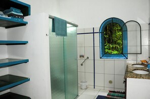Une salle de bain avec une douche à parois de verre, une fenêtre donnant sur une bambouseraie, un lavabo avec un plan de travail en granit et des étagères où sont pliées des serviettes.