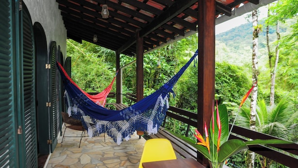 A covered outdoor seating area with a hammock, stone flooring, and a view of lush greenery. - Casa Caapuā, relax with comfort in nature. (Paraty)
