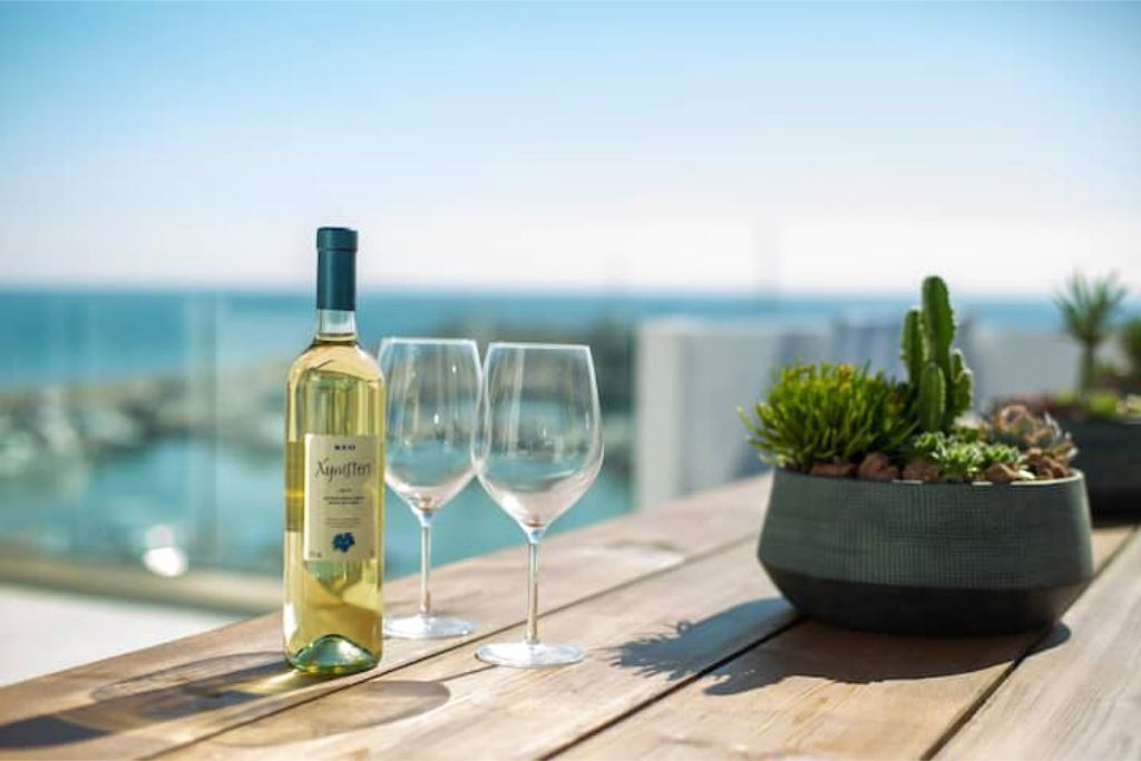 A bottle of wine and two glasses on a wooden table by a poolside with a view of the ocean.