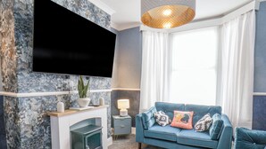 Uma sala de estar com televisão de tela plana na parede, um sofá azul com almofadas estampadas, uma mesinha de apoio com luminária e uma lareira.