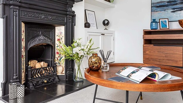 Una habitación con chimenea negra, una mesa redonda de madera con revistas, un jarrón con flores y un estante con adornos.