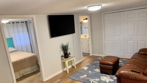 Ein kompakter Wohnraum mit Bett, Sofa, einem kleinen Tisch mit Pflanze, einem an der Wand befestigten Fernseher und einem durchgehenden Türspalt sichtbaren Badezimmer.