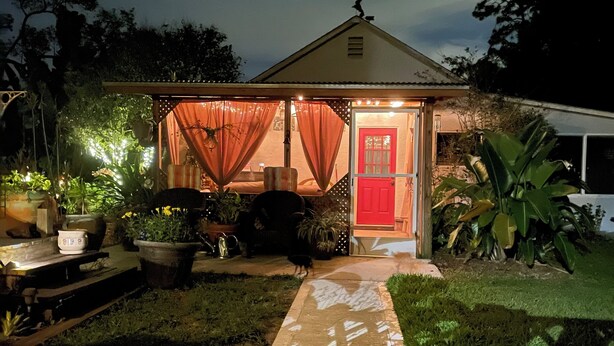Ein kleines Haus mit Veranda, einer roten Tür und Topfpflanzen.