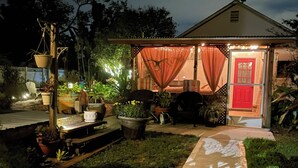 Ein kleines Haus mit roter Tür, umgeben von Topfpflanzen und Gartenmöbeln.