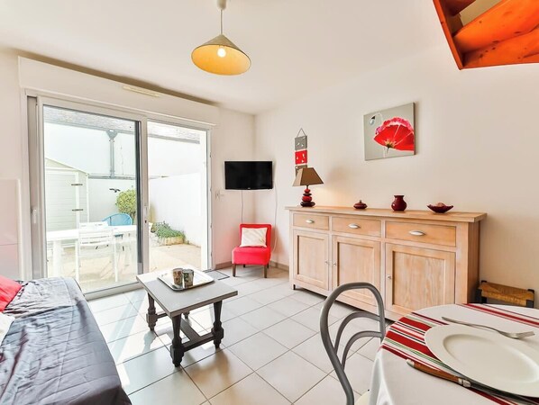 A modern living room with a sofa, a dining table with a red-striped tablecloth, a kitchen area with wooden cabinets, a red chair, and a television mounted on the wall.
