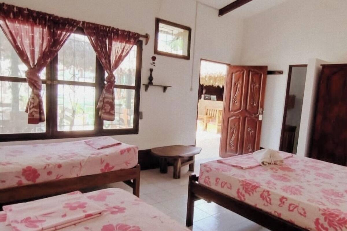 A room with two beds, a ceiling fan, wooden beams, a window with curtains, a small table, and a door leading to another room.